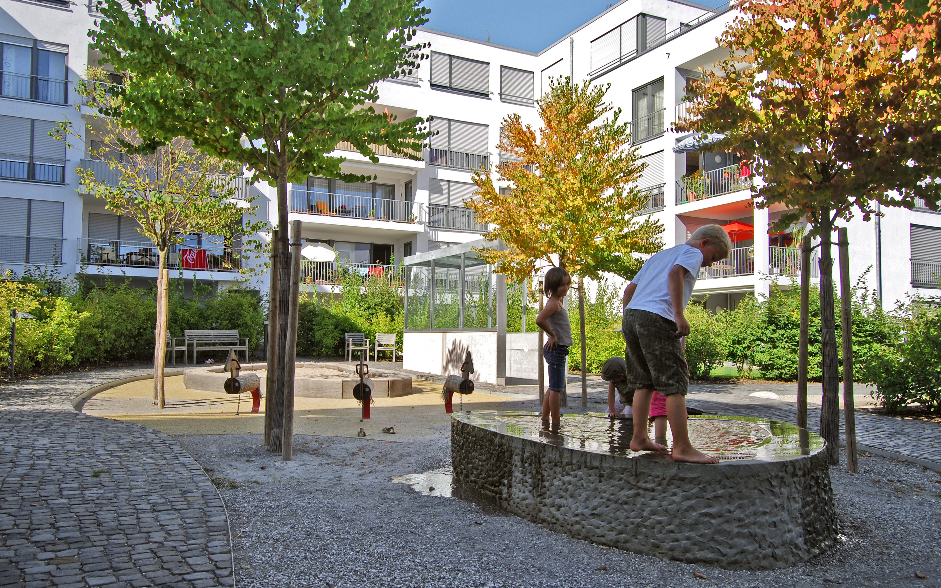 Baumbestandener Spielplatz mit Sandkasten, Wasserbecken und Reiterfiguren aus Holz